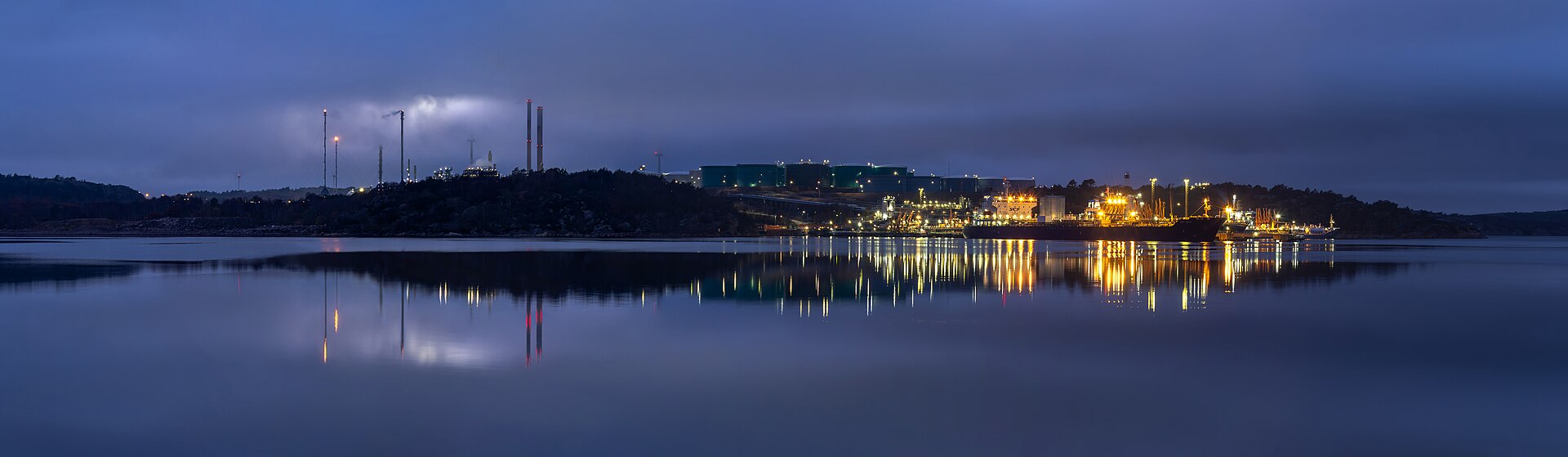 Vast oil refinery at blue hour, its lights reflecting across the water